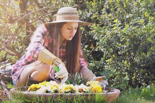 A young woman wearing a wide-brimmed straw hat, a red and black checkered shirt, and gardening gloves is kneeling on a grassy lawn in a back garden, tending to a flower bed filled with yellow and purple flowering plants. Behind her, there are dense green shrubs and trees, providing a natural backdrop. The garden features a mix of lush grass, soil, and decorative paving stones around the flower bed, with sunlight filtering through the foliage, indicating a bright, pleasant day. The scene captures a moment of outdoor gardening activity, illustrating typical garden maintenance and planting work that a gardening service in Surbiton, such as Hedge Trimming Surbiton, might support, emphasizing a well-maintained, leafy outdoor space ideal for leisure and horticultural care.