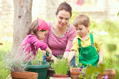 A woman engaged in gardening activities outdoors in a lush, green garden setting in Surbiton, with two young children assisting her. The garden features a well-maintained lawn with dense, vibrant grass, bordered by flower beds and potted plants in various shapes and sizes, including terracotta and plastic containers. In the background, tall trees and a wooden fence provide privacy and natural scenery. The woman is focused on tending to the plants, while the children, dressed in colorful clothing, help by potting or watering the plants. The outdoor space appears to be a private backyard or garden area, with natural sunlight illuminating the scene, suggesting a clear day. The overall environment emphasizes a healthy, maintained garden space with diverse greenery and flowering plants, reflecting professional gardening and outdoor maintenance services in the Surbiton area that support lush lawns, garden planting, and landscaping.