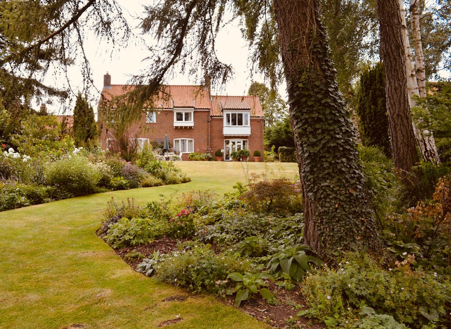 A view of a well-maintained garden in front of a red-brick residential building, with a gently sloping lawn bordered by flower beds filled with a variety of low-growing plants and shrubs. Several matu
