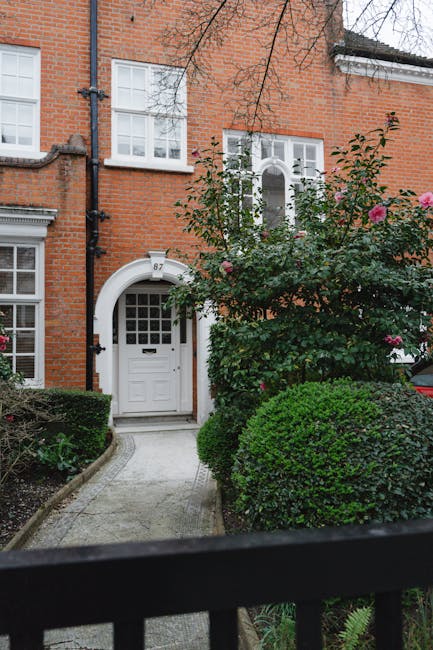 A view of a well-maintained garden in front of a red-brick residential building, with a gently sloping lawn bordered by flower beds filled with a variety of low-growing plants and shrubs. Several matu